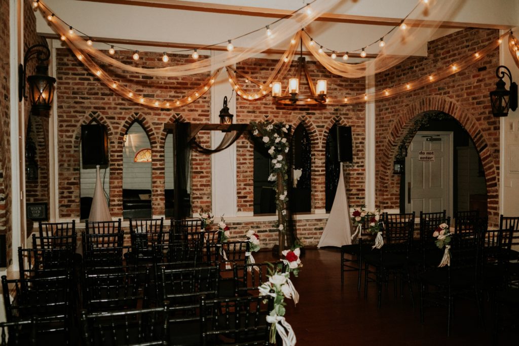 Indoor ceremony setup with wood arch and black chiffon drapery and white flowers in brick Historic Maxwell room