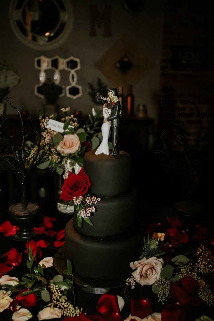 Gothic black fondant wedding cake with Bride of Frankenstein and Frankenstein's Monster cake topper surrounded by red and pink rose petals