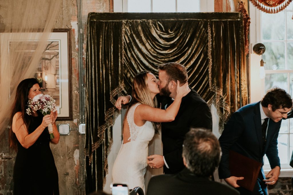 Bride and groom share first kiss in indoor wedding ceremony