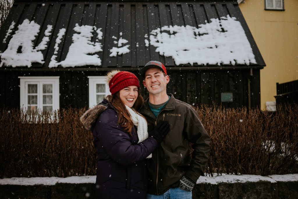 Couple smiles in front of black house in the snow on street in Reykjavík Iceland