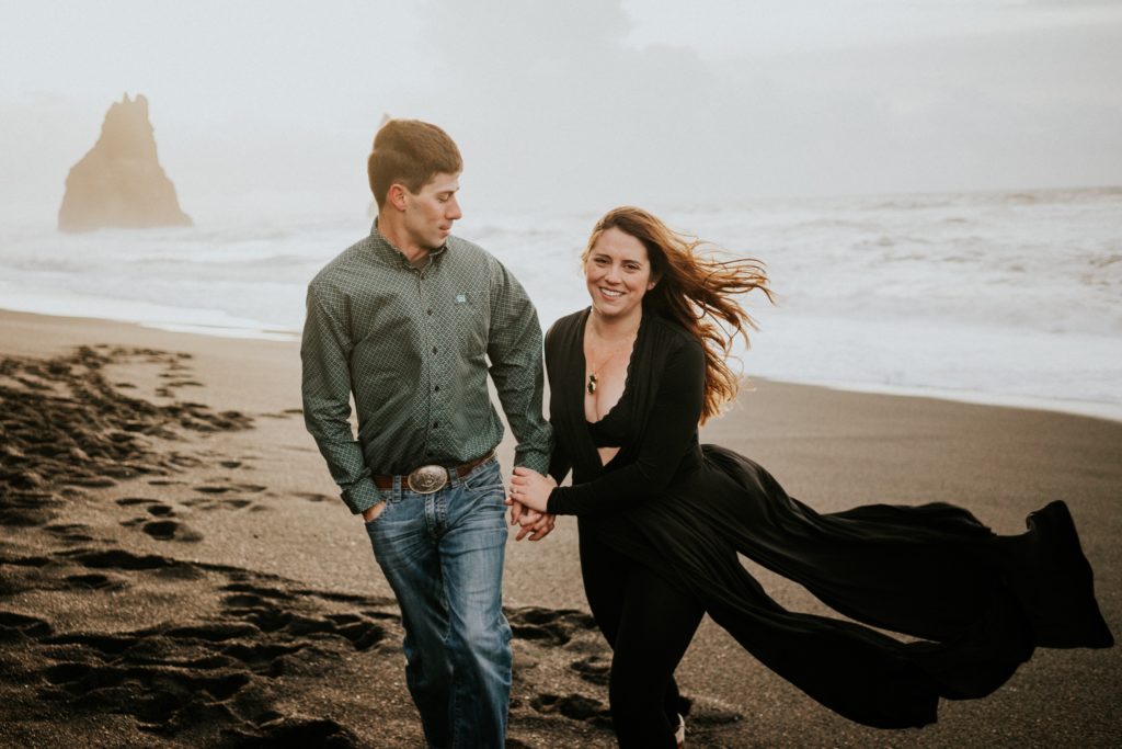 Couple hold hands smiling and walking with dress flowing behind them on black sand beach