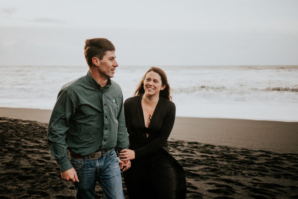 Couple hold hands smiling and walking on black sand beach