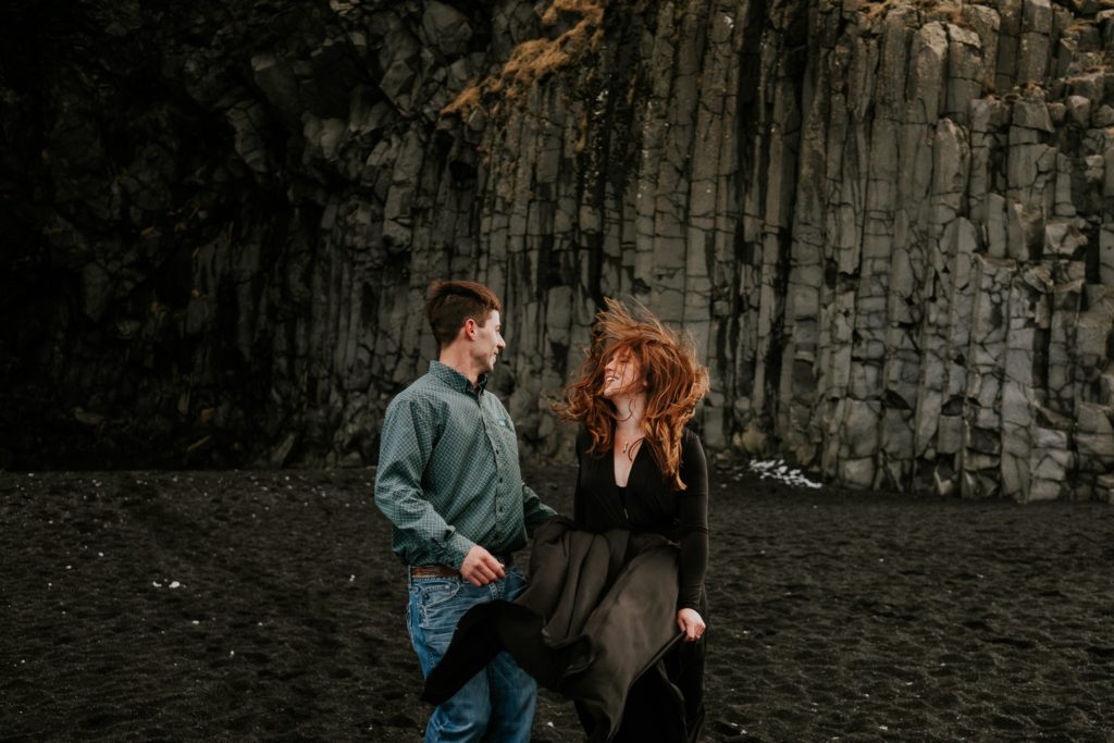 Couple's hair and clothes flow in the wind in front of basalt columns at Black Sand Beach