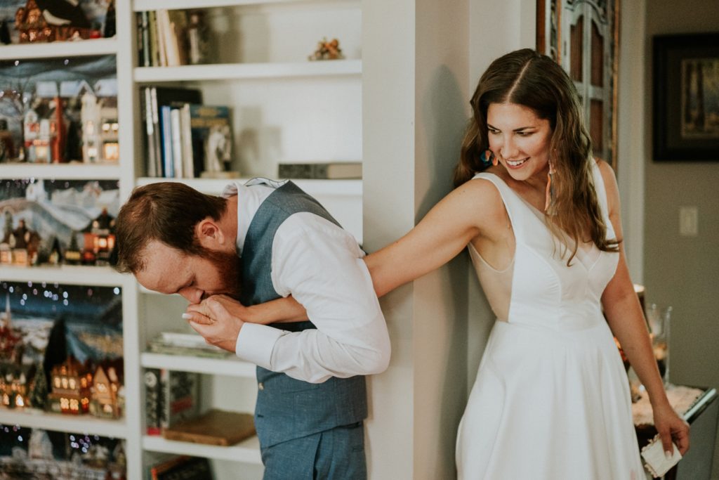 Groom kisses bride's hand as she peeks at him at home first touch