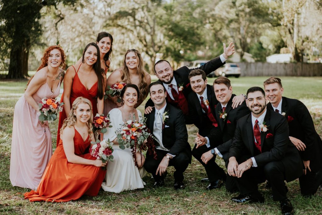 Wedding party in burgundy red and rust orange and black huddle together and smile