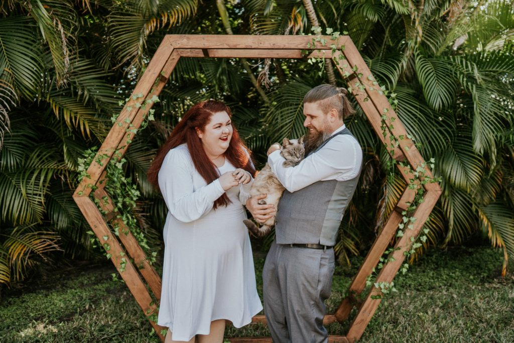 Redhead bride and groom hold cat under geometric hexagon arch in West Palm Beach FL backyard wedding