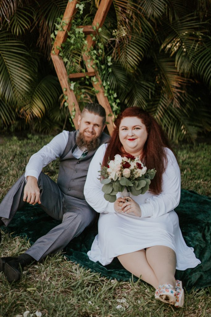 Redhead bride holding rose bouquet sits with groom in backyard wedding in West Palm Beach FL