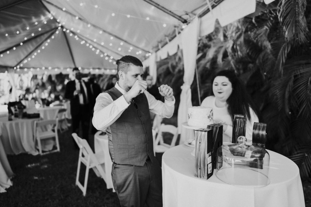 Groom raises fists in excitement eating cake FL backyard wedding in black and white