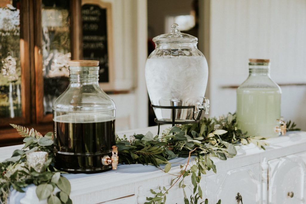 Soda, water, and lemonade jugs with greenery at rustic wedding bar