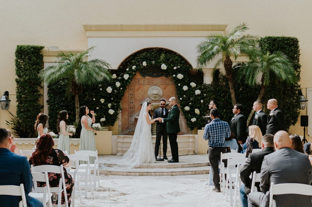 Benvenuto courtyard wedding ceremony fountain with white rose greenery wall
