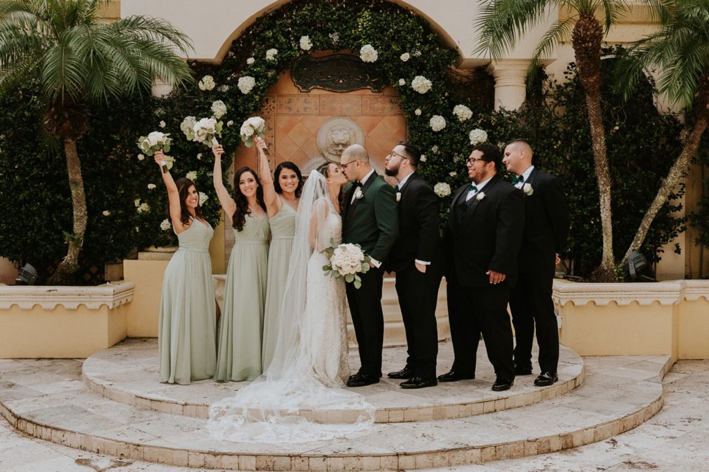 Benvenuto courtyard wedding party cheer as couple kiss in front of white rose greenery wall