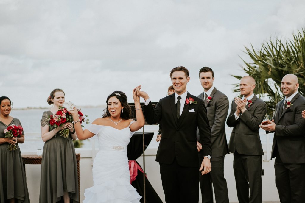 Bride and groom hold hands and cheer in wedding ceremony overlooking water
