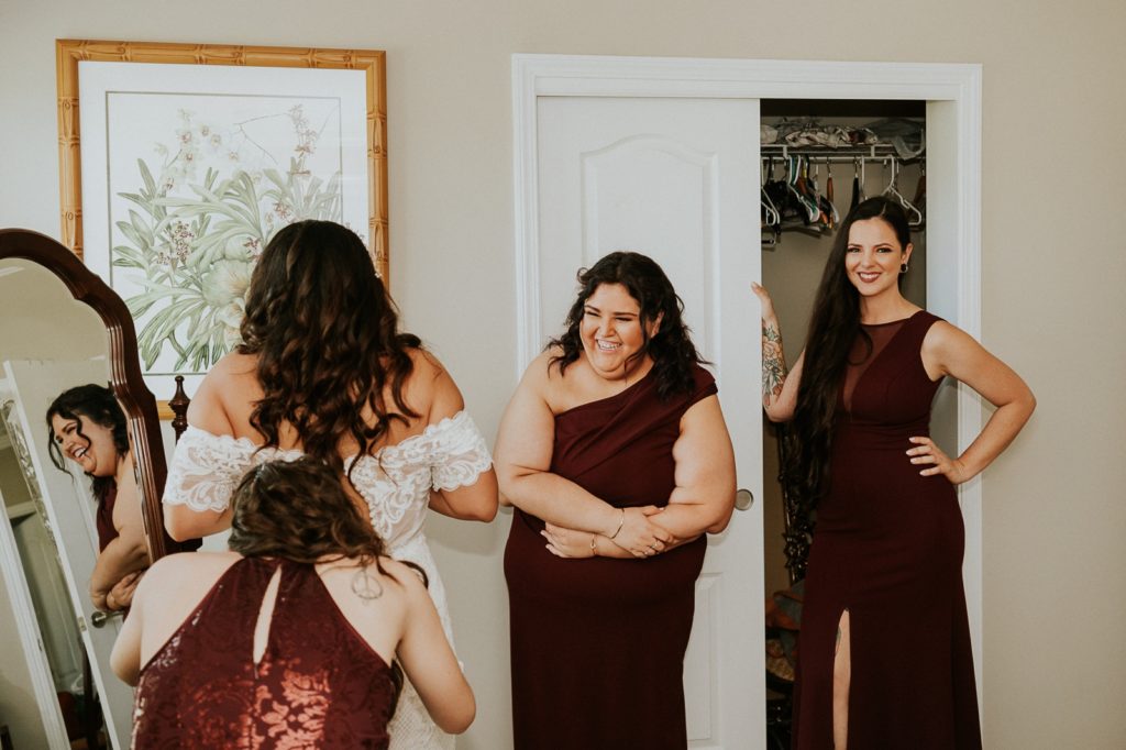 Bridesmaids in burgundy dresses help bride into dress