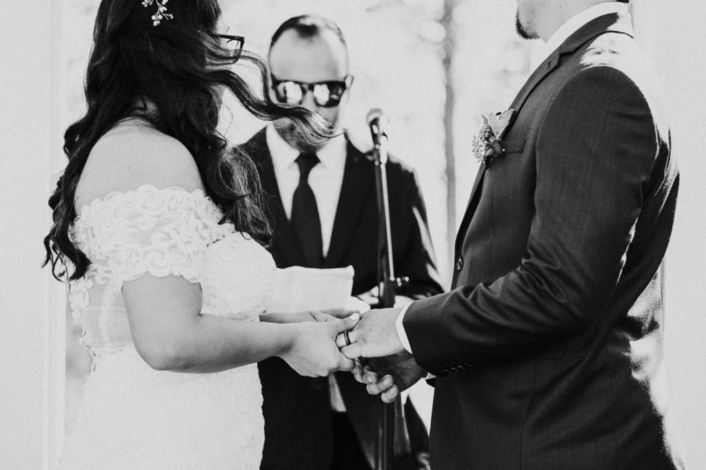 Black and white photo of bride and groom exchange rings during wedding ceremony