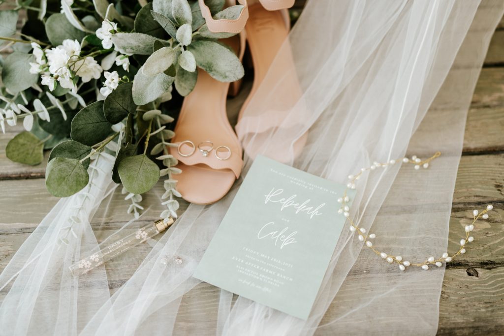 Wedding details flat lay with bouquet, shoes, perfume, and invitations on top of veil