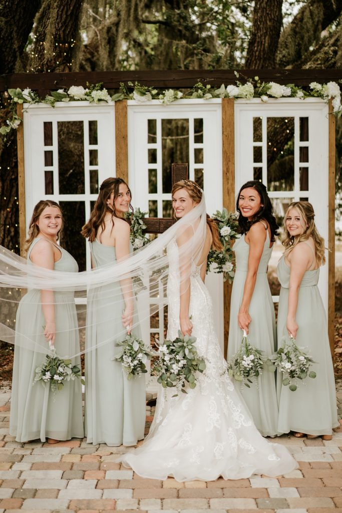 Bridal veil flows in wind in front of bridesmaids in sage green dresses