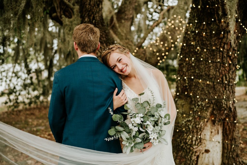 Bride holds grooms arm and smiles, veil flowing in the wind, with fairy lights on trees behind them at Ever After Farms Ranch