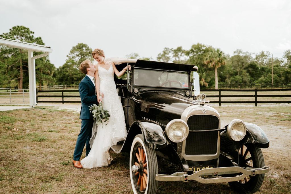 Groom supporting bride standing on black classic car at Ever After Farms Ranch wedding