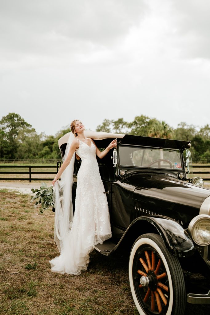 Bride standing on black classic car holding eucalyptus bouquet in the wind