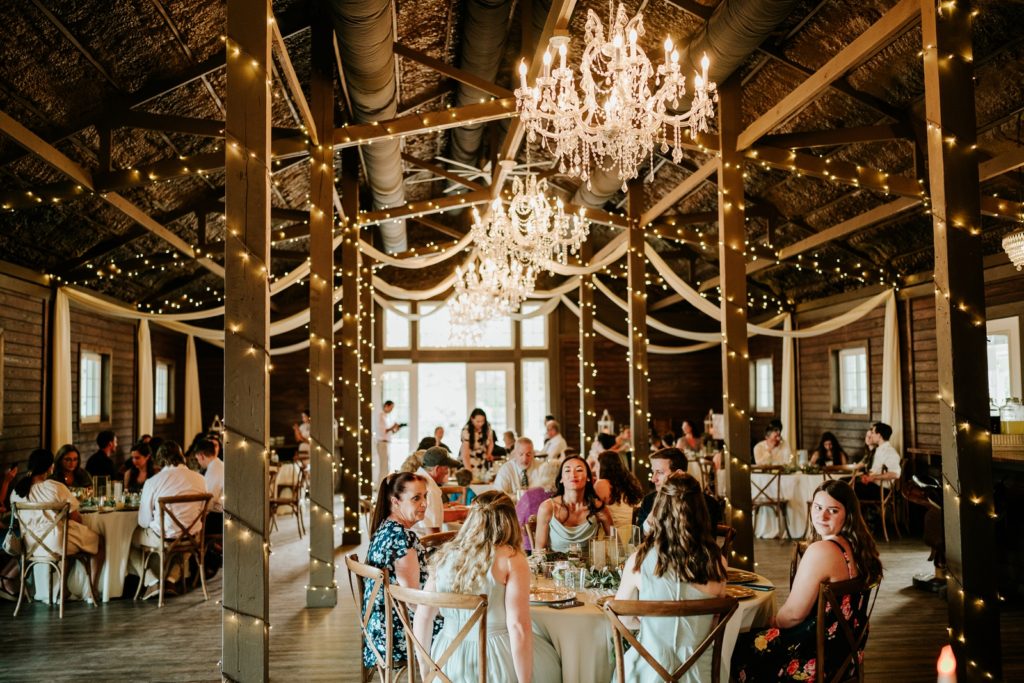 Guests sit for dinner in rustic wood barn with fairy lights and chandeliers at Ever After Farms Ranch wedding reception