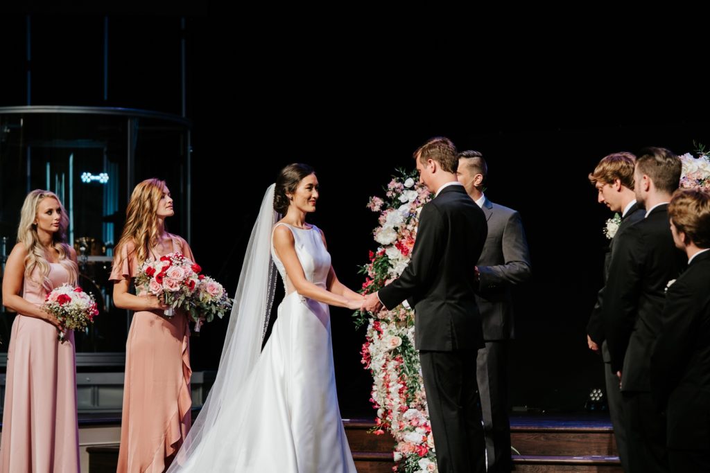 Bride and groom hold hands at indoor wedding ceremony between wedding party