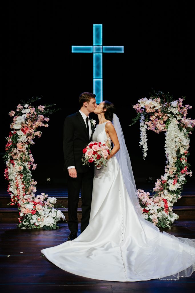Family Church Downtown wedding couple portrait kiss between floral arrangement arch under blue neon light cross