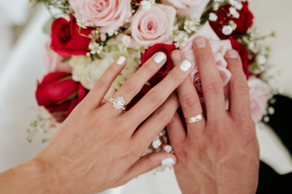 Wedding couple rest hands on top of pink and red rose bouquet showing off wedding rings and engagement ring