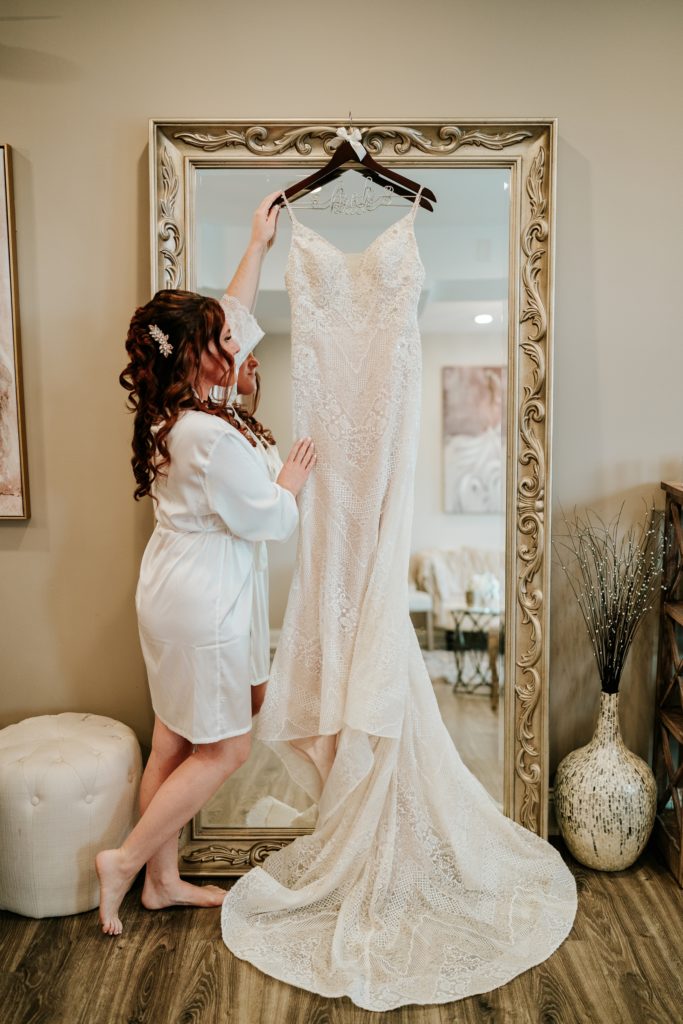 Bride hangs lace wedding dress on gold mirror in Pellegrino's Salon & Suite