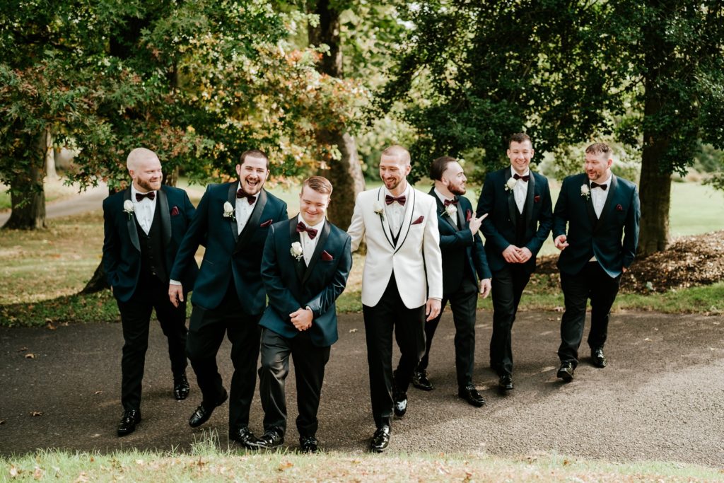 Groom in white suit walks with groomsmen in blue suits in fall wedding landscape