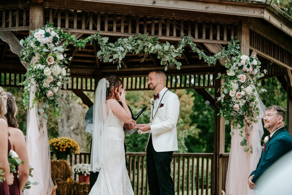 Bride wipes away a tear as groom smiles holding her hand under gazebo at Bensalen Township Country Club wedding ceremony