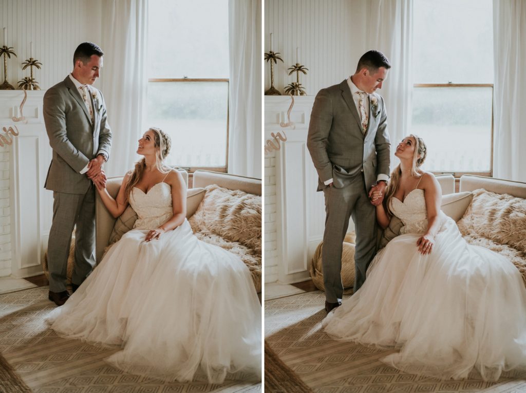 Bride sits on living room chair and groom stands beside her holding her hand