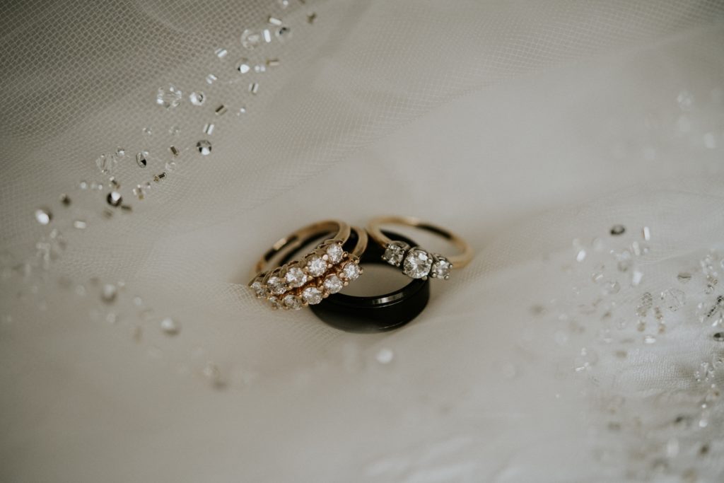 Women's gold and diamond engagement ring and wedding bands lay on top of black men's wedding band on top of a white veil with rhinestones