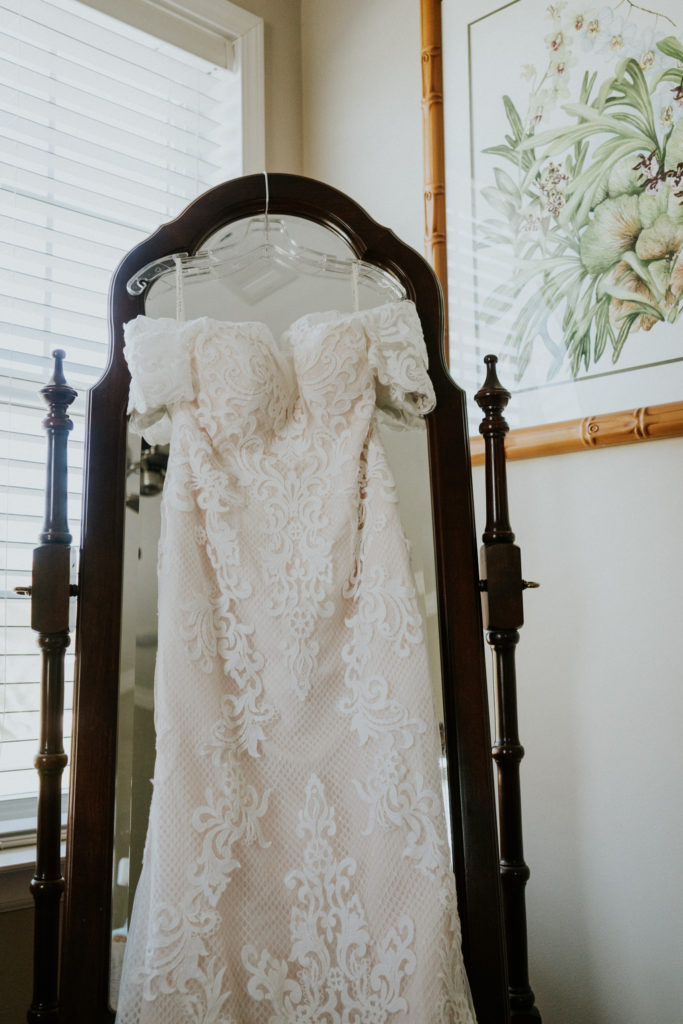 Fishnet lace appliqué off-the-shoulder wedding dress for Florida bride hanging on mirror at Kai Kai Farm Indiantown FL
