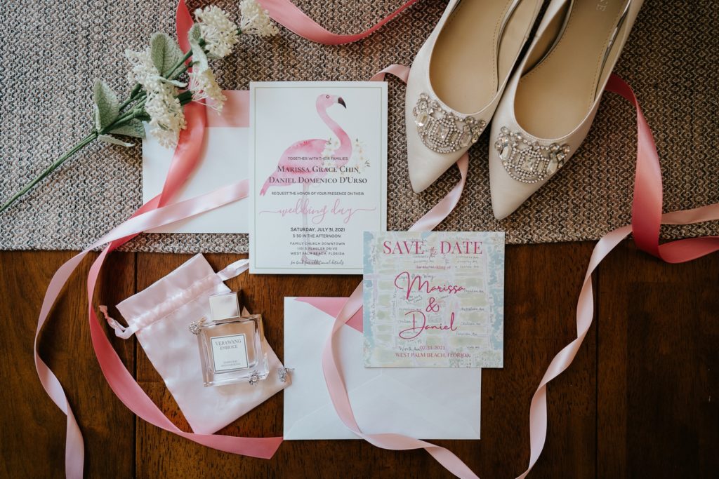 Wedding details flat lay with invitations, Vera Wang Embrace perfume, bejeweled shoes, and pink ribbon