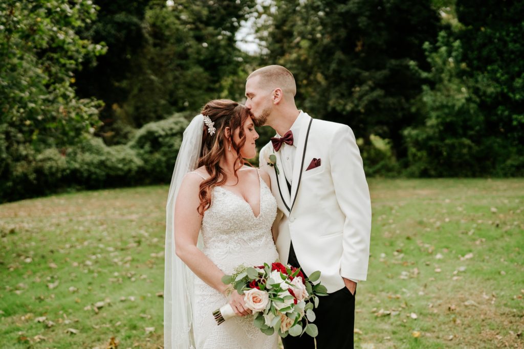 Groom kisses bride's forehead with lush greenery behind them