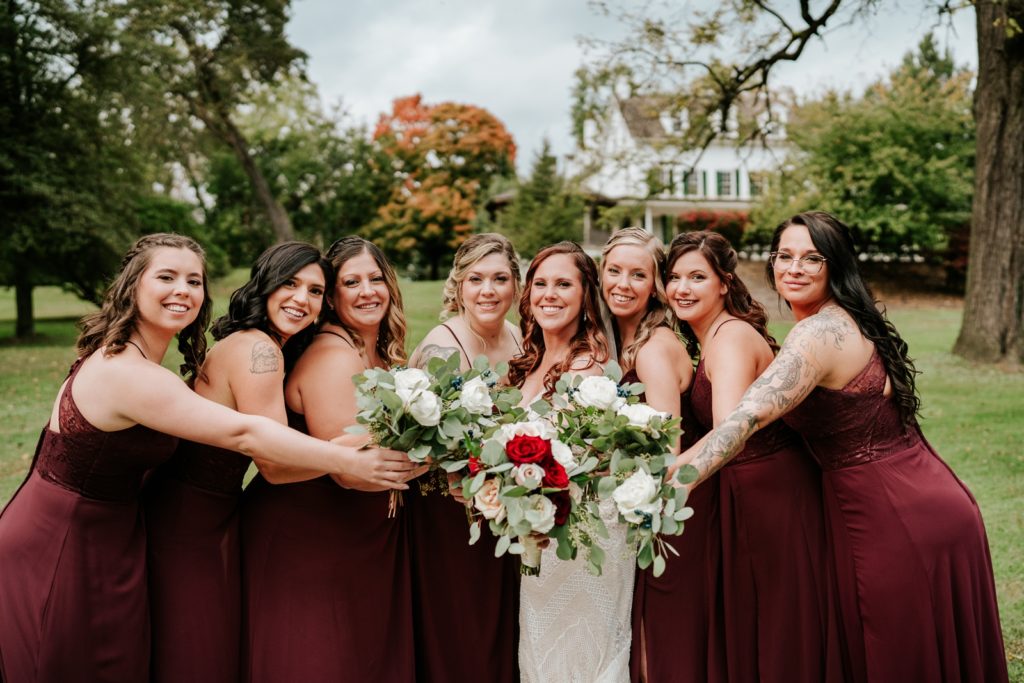 Bride and bridesmaids hold bouquets together in front of colorful fall trees at Bensalen Township Country Club