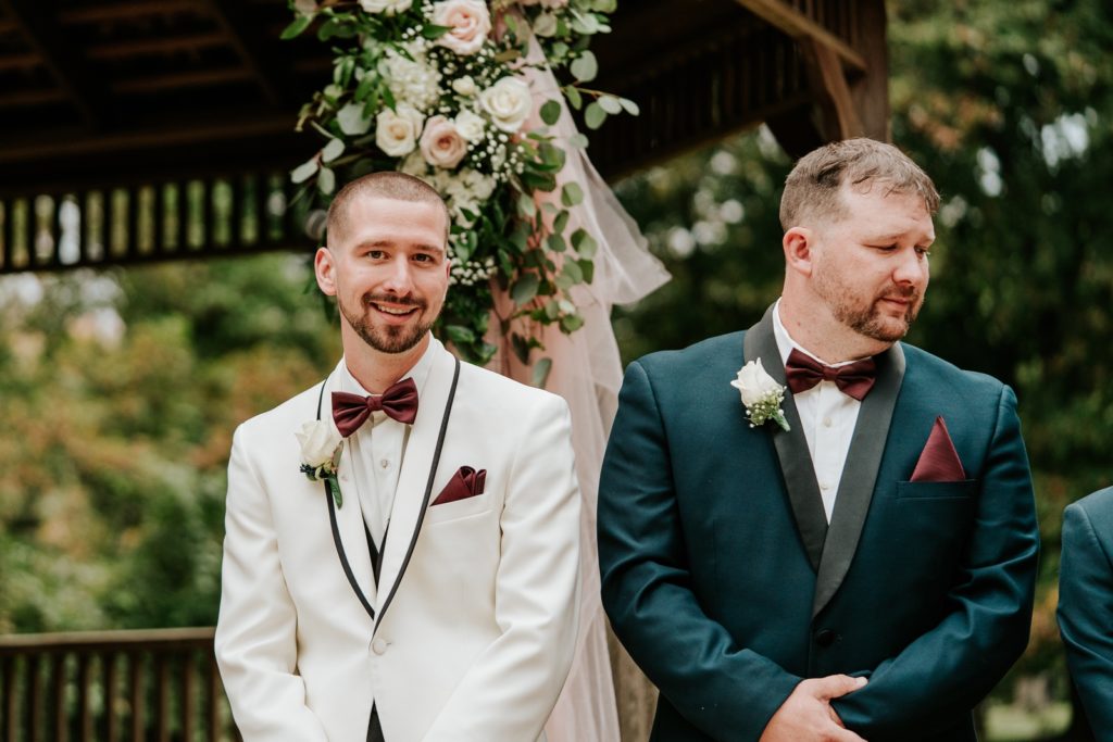 Groom smiles standing next to best man as he looks down the aisle to see his bride