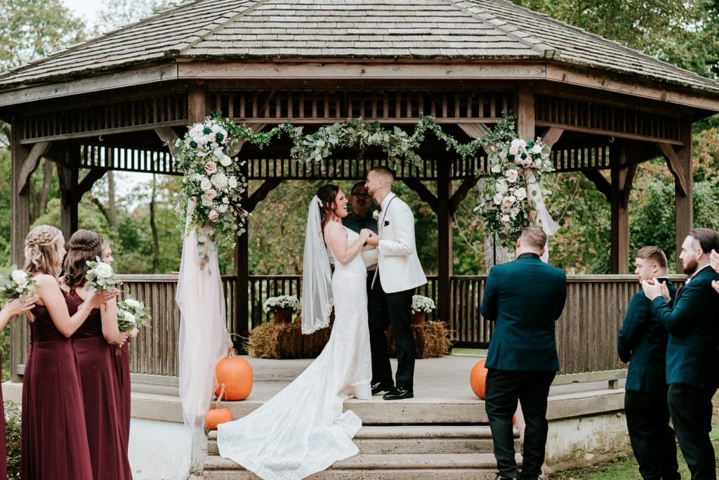 Bride and groom hold hands and laugh after first kiss under gazebo at Bensalen Township Country Club wedding ceremony