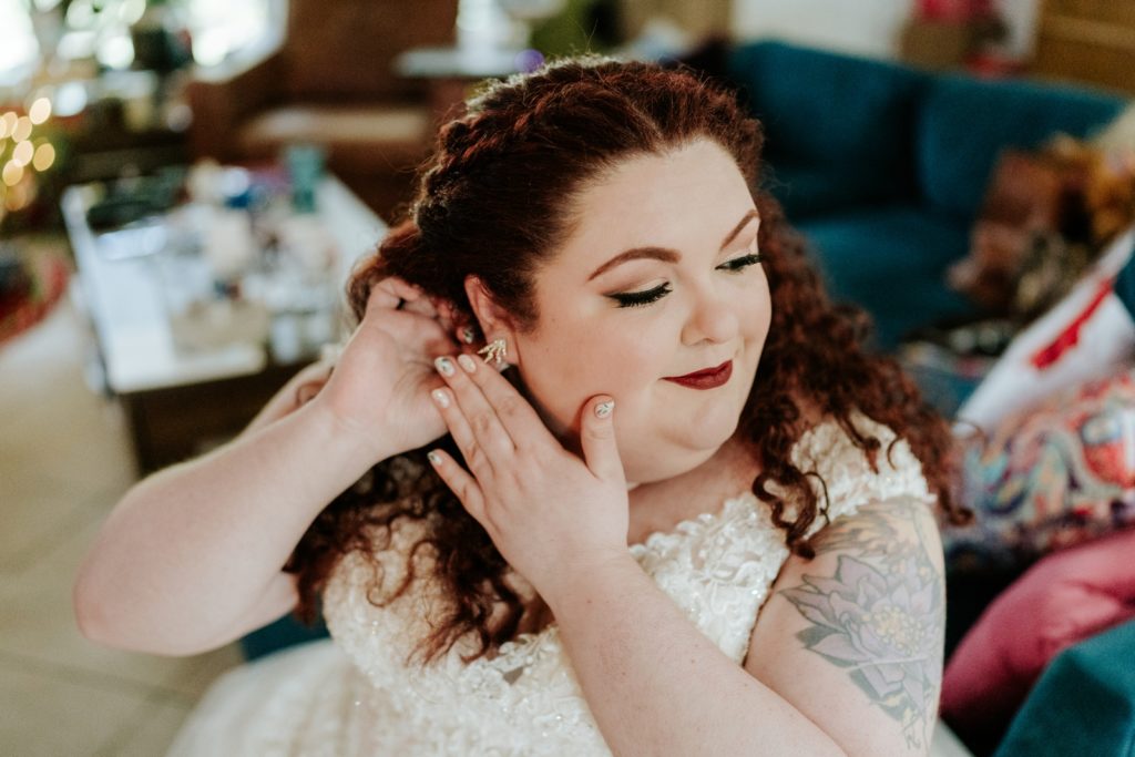 Bride with red curly hair and glamorous makeup adjusts earring, showing lotus flower tattoo on upper arm.