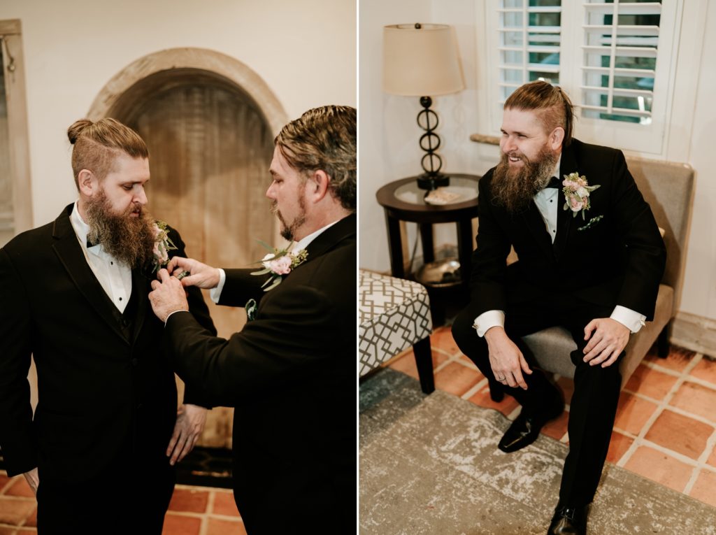 Best man adjusts groom's boutonniere and groom sits in chair wearing suit and laughing